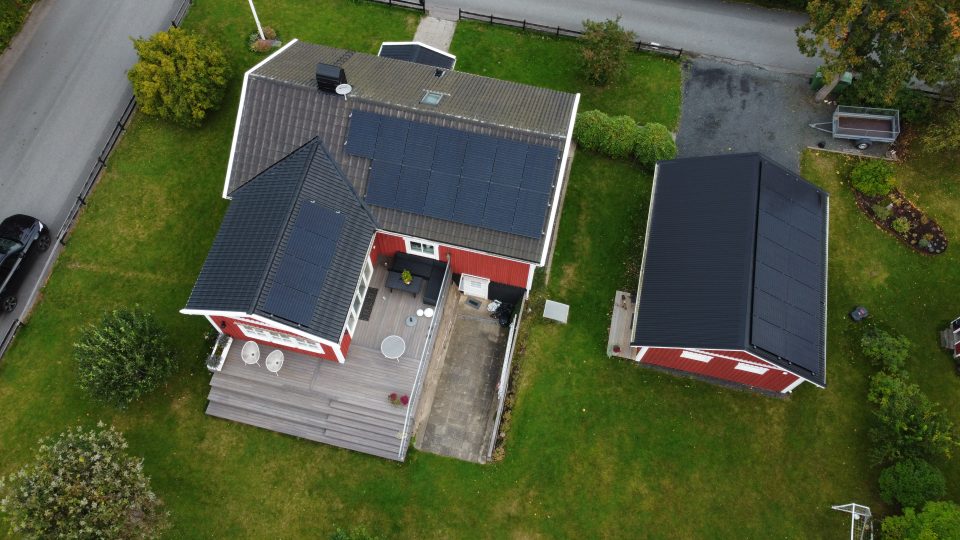 En översiktsbild av ett tak med solceller. Taket tillhör en röd villa med ett svart tak. Solcellerna är tydligt synliga och täcker en stor del av taket. Bilden är tagen rakt ovanifrån, vilket ger en tydlig vy över solcellerna och husets tak. Solcellerna är installerade i linjer och fångar upp solens strålar för att producera förnybar energi. Det röda huset och det svarta taket ger en kontrasterande färgkombination till bilden.