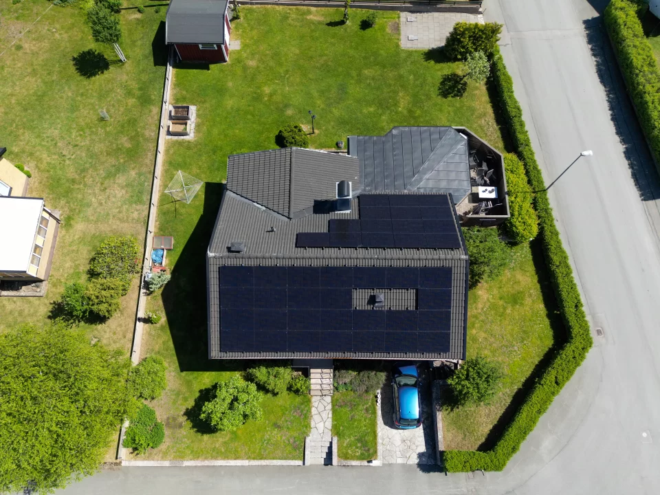 Bild på solceller installerade på ett tak, fotograferad rakt ovanifrån. I bilden syns trädgården och solpanelerna som sträcker sig över taket på båda sidor. Solpanelerna bidrar till hållbar energiproduktion och harmoniserar med den gröna omgivningen.
